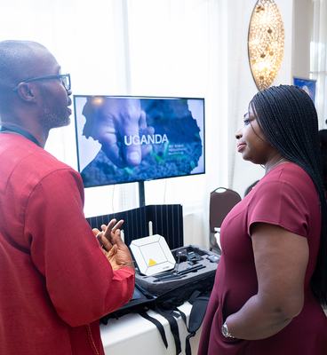 Two individuals stand beside a television screen in a well-lit room, engaged in conversation.