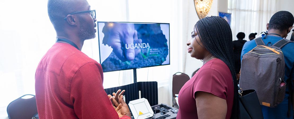 Two individuals stand beside a television screen in a well-lit room, engaged in conversation.