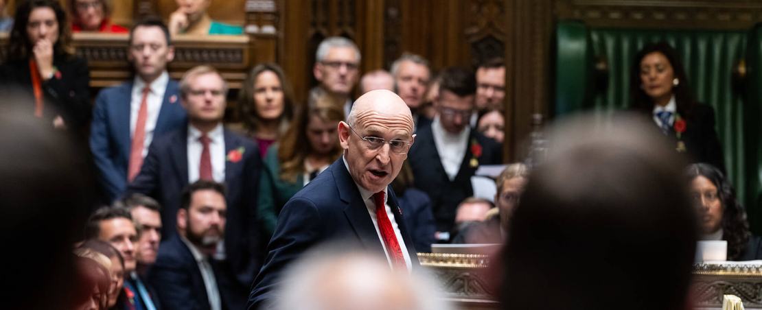 A man in a suit and tie delivers a speech in a parliamentary setting, addressing fellow members with confidence.