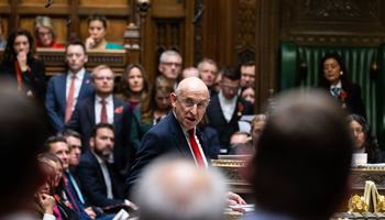 A man in a suit and tie delivers a speech in a parliamentary setting, addressing fellow members with confidence.