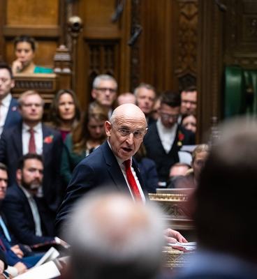 A man in a suit and tie delivers a speech in a parliamentary setting, addressing fellow members with confidence.