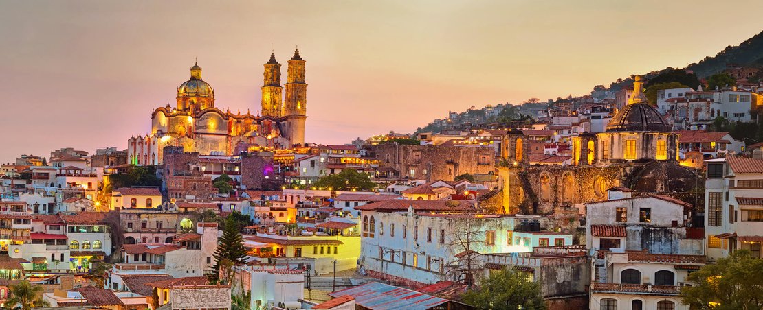 A city skyline at dusk featuring numerous buildings and a prominent church illuminated against the evening sky.