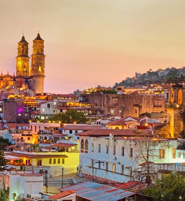 A city skyline at dusk featuring numerous buildings and a prominent church illuminated against the evening sky.