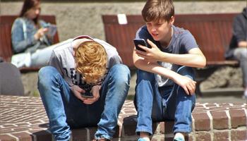Two teenagers seated outdoors, focused on their smartphones, embracing modern technology.