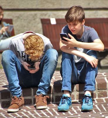 Two teenagers seated outdoors, focused on their smartphones, embracing modern technology.