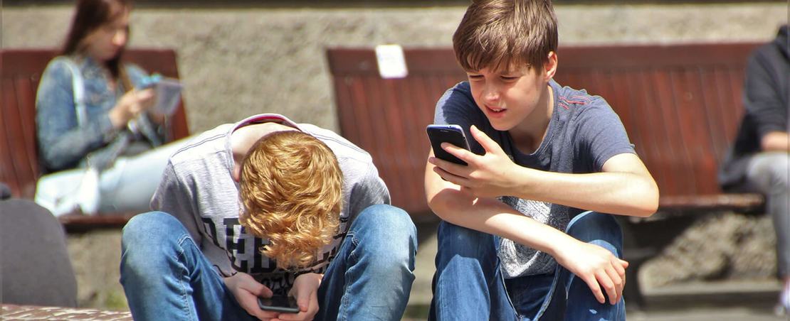 Two teenagers seated outdoors, focused on their smartphones, embracing modern technology.